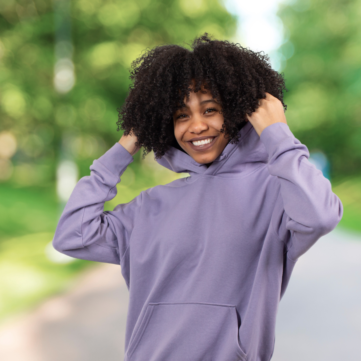 Smiling person in a light purple hoodie adjusting the hood while standing on a sunlit path surrounded by greenery. Featured on the Mind Rejuvenation Home page to reflect confidence, joy, and the uplifting power of nature in mental wellness.